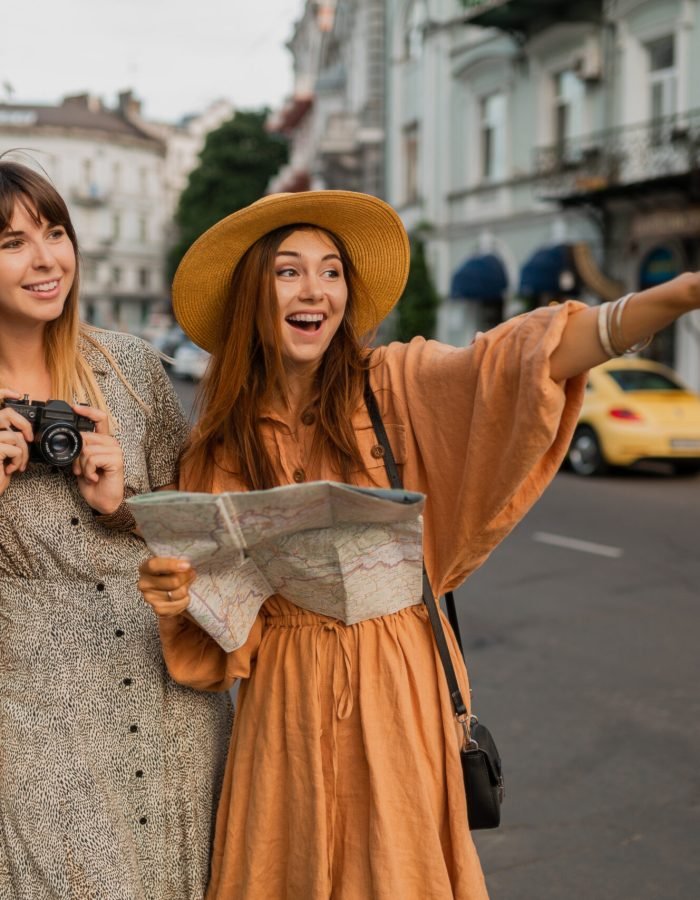 stylish young women traveling together in Europe dressed in spring trendy dresses and accessories smiling happy friends having fun taking photo on camera holding map